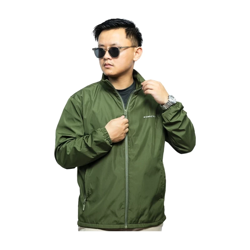 A male model wearing sunglasses looks off to the side while adjusting the collar of his olive green KTM CTY windbreaker, windcheater. The lightweight windguard jacket features a full-zip front, elastic cuffs, and a small white logo on the chest